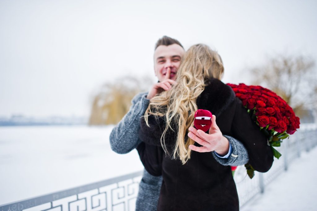 Jeune homme enlaçant son amoureuse et préparant la surprise d'une demande en mariage avec bague et bouquet de roses rouges.