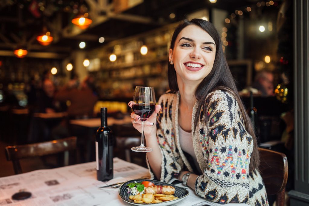 Jeune femme souriante attablée au restaurant, tenant une coupe de vin devant son repas.