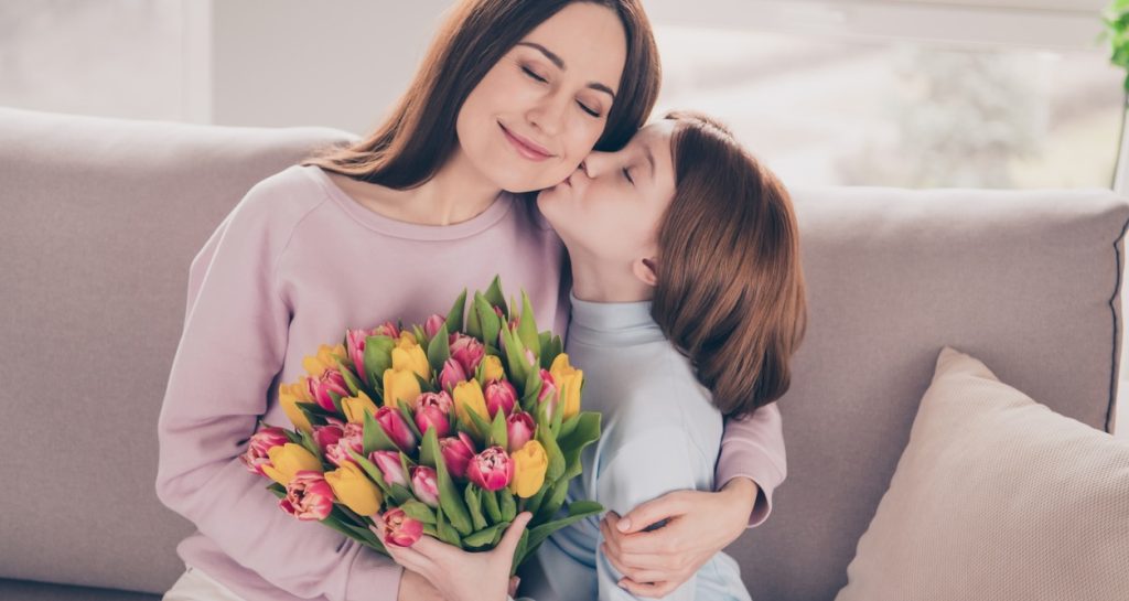 Fillette embrassant sa mère en lui offrant un bouquet de tulipes, toutes deux en tenue décontractée assises sur un canapé.